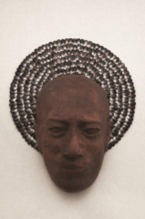 Fotografía de la escultura de un rostro humano hecho de barro, frente a un círculo compuesto de semillas negras dispuestas de forma radial