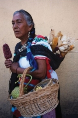 Fotografía de una mujer campesina de Xochimilco, sosteniendo una mazorca de maíz, cargando un atado de mazorcas en la espalda y soteniendo una canasta con productos vegetales